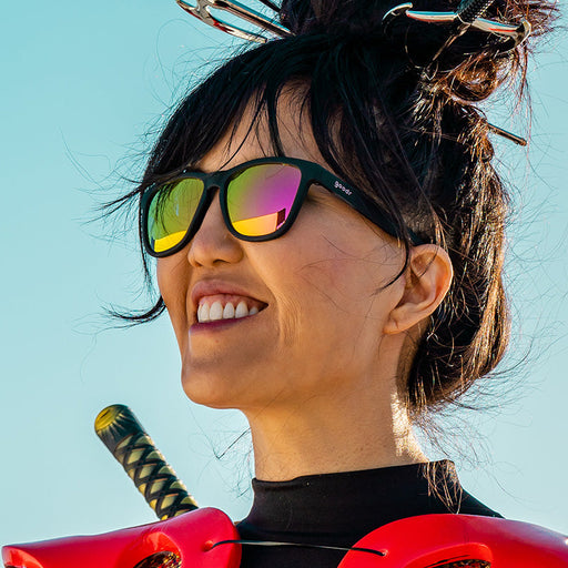 A woman wearing black sunglasses with hot pink reflective lenses wearing futuristic cosplay gear smiles into the distance.